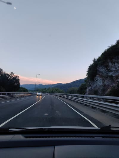 This image was taken from the interior of a vehicle on a highway at dusk, overlooking a mountainous landscape. The sky is a pale blue with a sliver of the moon visible in the upper left corner. The highway has multiple lanes and is bordered by a metal guardrail. Trees and a rocky cliff face are visible to the right of the highway. In the distance, hills are covered with trees and there are power lines visible on one of the hills. Two cars are on the highway, one white and one black. The road surface appears wet, reflecting the headlights of the cars. The dashboard of the vehicle is visible in the foreground. The image does not depict any specific location within the city of Monastyr, Russia.
