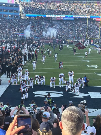A large crowd fills stadium seats at a football game, the field displaying white yard lines. Smoke rises from the crowd. Two teams, one in white and the other in black uniforms, line up along the edge of the field. People on the sidelines take pictures. A scoreboard shows “SEAHAWKS VS PATRIOTS” and “SEATTLE SEAHAWKS.” Numbers such as "48," "15," and "25" are visible on the players' jerseys. Logos like "Titleist" and "POLO" appear on caps and other merchandise.