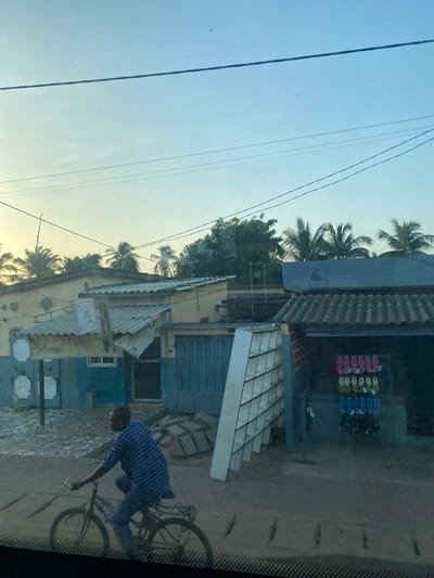 A street scene in Aného, Togo, features a man wearing a blue striped shirt riding a bicycle. Buildings are visible along the street, including a shop displaying footwear for sale. The weather appears to be clear and sunny, and palm trees are seen in the background. Overhead power lines are present. The image seems to be taken from inside a vehicle, looking out onto the street.