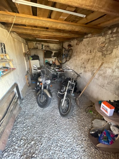 The image depicts the interior of a dimly lit, rustic garage or storage space, likely located in Znamensk, Russia. The walls are constructed of rough, aged plaster and exposed brick, with visible signs of wear and tear. The ceiling is supported by rough-hewn wooden beams, with a fluorescent light fixture attached. The floor is covered in coarse gravel.
In the center of the space, two motorcycles are parked. The motorcycle on the left is a cruiser-style bike with a silver-grey finish, a large windshield, and chrome accents. The motorcycle on the right appears to be a sportbike or custom motorcycle, with exposed engine components and a more aggressive stance.
Various items are scattered around the garage, suggesting it is a working or storage area. To the left, a wooden shelf unit holds tools and other miscellaneous objects. In the background, shelves are lined with storage containers and bags. A bicycle is visible mounted on the wall in the upper right portion of the image, and a shovel leans against the right wall. On a circular table to the right, a red and white battery pack is prominently displayed, along with other items. The overall impression is one of a cluttered, functional space used for vehicle maintenance and storage.