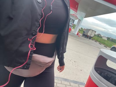 The image shows a person at a gas station, likely in Molokovo, Russia, based on the surrounding environment. The person is wearing black clothing and a light-colored hip bag. Pink headphones are visible. In the background, there is a gas station canopy, gas pumps marked with "EVOLUTION EVO" and the number "1", and a truck driving on a road. Houses are seen in the distance under a cloudy sky. A red and gray trash bin is also visible.