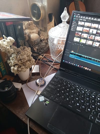 This is a close-up, eye-level shot of a dark wooden desk surface, cluttered with various objects. In the foreground, a black laptop is open, its screen displaying video editing software with a timeline and thumbnail previews of video clips. The laptop's keyboard and trackpad are visible, along with Intel Core i7 and NVIDIA GeForce stickers.

To the left of the laptop, a black camera lens is positioned, and next to it, a small white box. Further left, a bouquet of dried white flowers is arranged in a small white vase. Behind the flowers, a stack of books is visible. The spines of these books show titles and author names, including "Yuvall Noah Harari," "Abdullah Hehamahuwa," and "1984."

To the right of the flowers and books, a small, seated Buddha statue is present. Behind the Buddha statue and to the right, a large, ornate, clear glass candy jar filled with amber-colored liquid is prominent. Further back, a round clock with a dark face and gold trim is mounted on a wall or background structure. A portion of an acoustic guitar is visible in the upper right corner.

A lighter, possibly a Zippo, lies near the center of the desk. A few cables snake across the surface, connecting to the laptop and other unseen devices. A round, metallic pin with an image is also visible on a piece of white paper. The overall impression is of a workspace with personal items, suggesting creative activity. The lighting suggests it is daytime, likely indoors.
