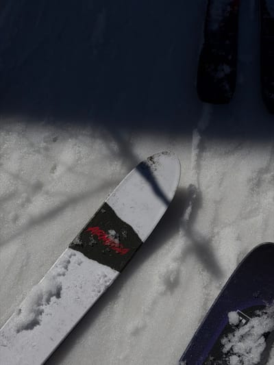 The image depicts three skis resting on granular snow under bright sunlight, casting distinct shadows. The scene is located in Saint-Chaffrey, France.

In the foreground, angled from the bottom left towards the center, is a white ski with a prominent black section featuring red, stylized text that appears to read "ARMADA." Snow adheres to the surface of this ski. To the bottom right, a portion of a dark-colored ski, possibly purple or black, is visible, also with snow accumulation. In the upper right quadrant, mostly obscured by shadow, are the tips of two dark objects, likely ski bases or boots, resting upright. A sharp shadow line traverses horizontally across the middle of the frame, separating the brighter, sunlit snow in the lower half from a significantly darker, shadowed area in the upper half. Multiple elongated shadows, consistent with those cast by trees or poles, are visible across the snow. No individuals are present or engaged in any activity.