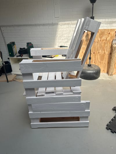 In Lisse, Netherlands, a white painted pallet chair is positioned in what appears to be a workshop or garage. The chair is made from disassembled and repurposed wooden pallets, with the backrest angled upwards. The chair is partially painted white, with some areas of raw wood visible, suggesting it is either a work in progress or a rustic design. The floor is a concrete surface. In the background, a white brick wall is visible, along with various items including two green toolboxes, coiled cables, a white cabinet, a large black punching bag, and sheets of plywood. Sunlight streams in from an unseen window, casting shadows across the scene.