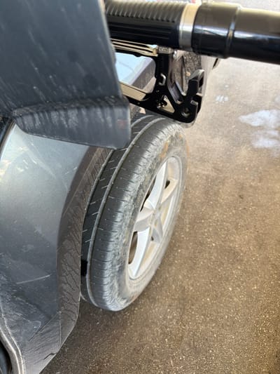 In Meljine, Montenegro, a close-up shot focuses on the side of a dark-colored vehicle, specifically its rear tire and part of the fender. A black fuel nozzle is inserted into the fuel door of the car. The tire is moderately worn and shows some dirt. The surrounding ground appears to be asphalt or concrete, with a light scattering of what might be sand or dirt. The lighting suggests an outdoor, daytime scene.