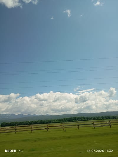 An exterior, medium-shot photograph captures a wide, grassy field bordered by a wooden fence, with a dense line of green trees behind it. In the distance, a range of muted, blue-gray mountains forms the horizon. Above the trees, a band of white, puffy clouds stretches across a bright blue sky. Several dark, horizontal power lines traverse the sky. The bottom left corner of the image displays the text "REDMI 13C," and the bottom right corner shows the date and time, "16.07.2026 11:32." The scene is set in Galbai, Russia.