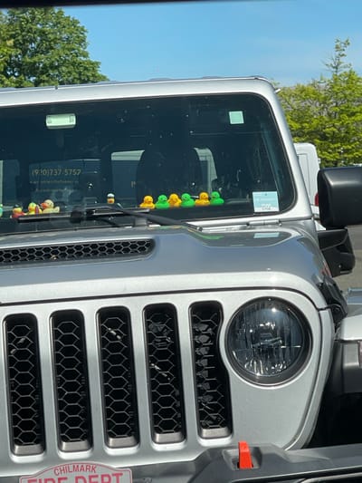 A silver Jeep Wrangler is parked outdoors on a sunny day. The Jeep's windshield is visible, and on the dashboard, there are several small rubber ducks lined up. A sticker with "myfp 0706" is affixed to the passenger side of the windshield. The Jeep's grille, headlights, and part of the hood are also in view. In the background, trees and a blue sky are present. The license plate frame on the front bumper reads "CHILMARK FIRE DEPT". The scene is likely in Edgartown, Massachusetts, as indicated by the "Chilmark Fire Dept" detail.