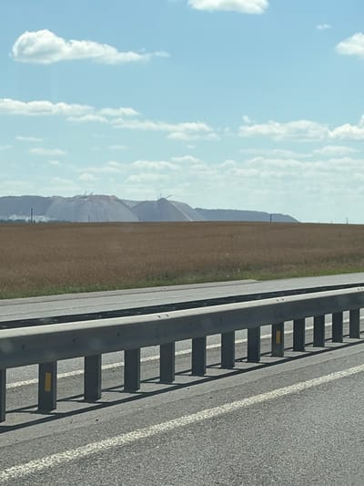 The image depicts a wide, flat landscape under a bright blue sky with scattered white clouds. In the foreground, a paved road with white lane markings is visible, separated by a metallic guardrail. The guardrail consists of vertical posts supporting a horizontal beam, with yellow rectangular reflectors attached to some of the posts. Beyond the road and guardrail lies a field of dry, golden-brown grass or crops. In the distance, a range of large, pale mounds dominates the horizon. These mounds appear to be artificial, possibly waste piles from mining or industrial activity, with several cranes or heavy machinery visible on their slopes. The overall impression is one of an industrial or agricultural area with a wide-open sky. The location is identified as being within the Belitski Sel'ski Sovet, Belarus.