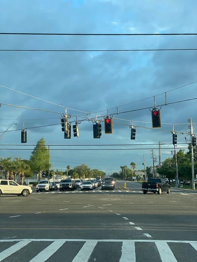 The image depicts a street scene in Largo, Florida, United States of America. The view is likely from inside a vehicle. The street is lined with multiple vehicles stopped in traffic. Overhead, several traffic lights are visible, all displaying red arrows. The sky is overcast with dark clouds. Vegetation and buildings line the roadside. Crosswalk markings are visible on the road. Power lines are strung across the sky.