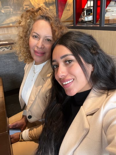 Two women are posing closely together. The woman on the left has curly blonde hair and wears a white top and a beige jacket. The woman on the right has long black hair and wears a black top and a beige jacket. Both are smiling at the camera. In the background, part of a staircase and some furniture are visible.