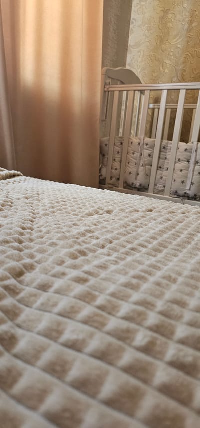A close-up, low-angle shot captures a quilted, beige blanket or bedspread in the foreground, with a pattern of small, raised squares. In the background, partially obscured by sheer, peach-colored curtains, is a white crib with decorative padded bumpers featuring a pattern of gray stars. The wall behind the crib appears to have textured, patterned wallpaper. The setting is likely a bedroom, possibly a nursery, and given the context of a crib, it suggests a residential interior. The image does not provide any specific clues to pinpoint its location within the city of Овощной, Russia.