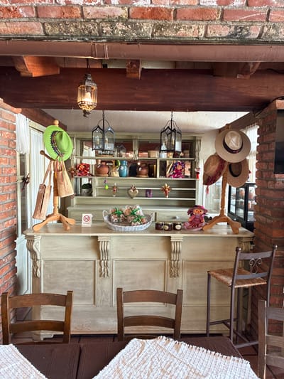 The image depicts a partially covered outdoor display in Tepeji de Ocampo, Mexico. The display features shelves with various artisan goods including pottery, hats, and dolls. A light fixture is visible on the left, near brick and wooden architectural elements. Additional hats are hung on wooden rack. Two wooden chairs sit to the right, partially obscured by a white textile. The view is from below, looking upwards into a space framed by brick walls and overhead structures.