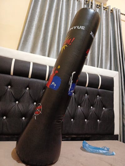 This media file shows a black inflatable punching bag standing upright on a bed. The punching bag is leaning against a dark upholstered headboard with white vertical stripes. Behind the bed, there are silver-grey curtains and a portion of a white air conditioning unit.

The punching bag has several cartoon-like graphics printed on it. One graphic depicts a red monster with a blue, round-faced monster below it, and a yellow blob above them. Another graphic shows a target with the words "GAME ON!" written in red and white. The word "INYUE" is printed in large white letters near the top of the punching bag, and the letters "XIN" are also visible.

A crumpled blue plastic bag lies on the bed in the foreground, to the right of the punching bag. The lighting suggests it is likely nighttime or indoors. The overall scene appears to be a bedroom in Payacut, Indonesia. No people are visible. The activity taking place is not evident, but the presence of the punching bag suggests it is for exercise or recreation.