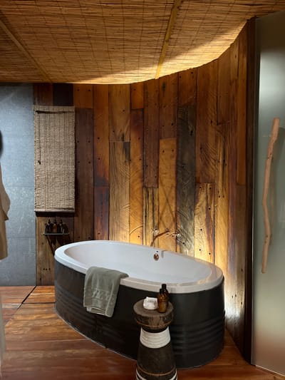 The image depicts a bathroom interior, possibly in Livingstone, Zambia, given the use of natural materials in a rustic design. The room features walls constructed from horizontal wooden planks, creating a textured backdrop. A curved wall covered in a woven bamboo-like material adds a tropical element to the design. A bathtub, encased in what appears to be a repurposed metal barrel painted black, is positioned in the corner of the room. A towel is draped over the edge of the tub. A small wooden side table next to the tub holds a bottle and some other smaller items. A woven basket and a small metal wine rack holding bottles are visible on the floor. The floor itself is tiled in a light gray color. A decorative wooden branch is mounted on the wall near the ceiling.