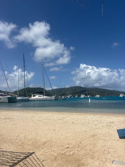 The scene depicts a sunny daytime view of a harbor in Spanish Town, British Virgin Islands. The foreground consists of a light-colored sandy beach, with a dark shadow of a potential fence or railing cast on the sand in the lower-left quadrant. A partial dark blue object is visible on the sand in the lower-right. The midground features clear, light blue-green water transitioning to deeper blue. Numerous boats are present: several sailboats and catamarans are moored closer to the shore, including a white catamaran labeled "Sweet Chariot." Further out in the bay, approximately twenty additional vessels, primarily powerboats and some sailboats, are anchored or moored. A white cylindrical buoy floats in the center-right water. The background reveals lush, green, undulating hills or mountains under a bright blue sky with scattered white cumulus clouds. No individuals are visible.