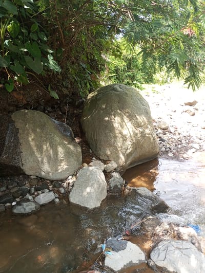 A natural outdoor scene in Cibeureum, Indonesia, captured under bright sunlight. The primary subjects are a murky brown stream flowing from upper left to lower right, flanked by dense vegetation and numerous rocks. Two prominent, large, grey boulders are centrally positioned, partially submerged or adjacent to the water, casting distinct shadows. The left bank is characterized by a steep incline covered in thick green foliage, including broad-leaved plants. The upper background displays dense green tree canopies. Within the stream, particularly in the lower right quadrant, discarded human refuse, including various colored plastic fragments, is visible amongst smaller rocks. No human subjects or structures are present.