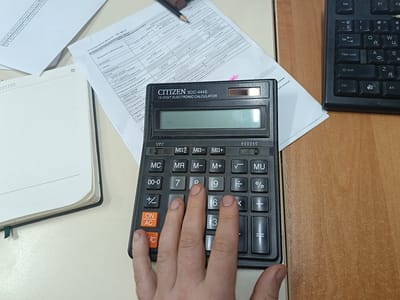 The image shows a close-up, top-down view of a desk with a calculator, papers, and a notebook. A person's hand is using the calculator, with fingers pressing buttons. The calculator is a black CITIZEN SDC-444S 12-digit electronic calculator. Papers with text and tables are spread out on the desk, one with a pencil on it. A notebook lies open to the left. A keyboard is partially visible on the right. The desk appears to be made of wood. The scene is not specific to the city of Zaslaŭje, Belarus; it is a generic office or study desk setting.