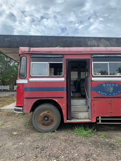 An aging, red bus with a blue stripe is parked on a gravel lot. The bus has its door open, revealing a set of metal stairs leading into the interior. The bus has several windows, some with visible reflections of trees and sky. A circular emblem with a lion and writing is visible on the side of the bus. A concrete overhang extends over the top of the bus, suggesting it might be at a bus station or shelter. The environment appears to be outdoors, with some sparse green vegetation growing in the gravel. The sky is cloudy. The image does not contain any people or specific actions occurring. The scene is static and appears to be a view of the bus from the side.