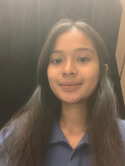 A sideways portrait photograph, rotated 90 degrees counter-clockwise, depicts a young woman with long, dark hair, smiling gently and looking directly at the camera. The subject, likely in Samarinda, Indonesia, has a light to medium complexion, dark eyes, a small nose, and full lips. Her hair, which appears to have some subtle lighter streaks, flows around her and across the frame. She is wearing a dark blue top with a visible V-neckline. The background is largely dark and indistinct, suggesting an indoor setting with soft, ambient lighting illuminating her face from the left. The image exhibits a noticeable graininess, consistent with low-light conditions or a specific photographic aesthetic. The woman's expression is calm and friendly. No specific activity or event is taking place beyond posing for the photo, and no text is visible.