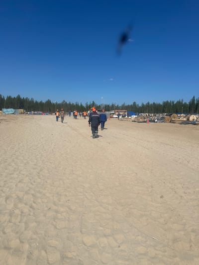 The image depicts a group of people, appearing to be construction workers, walking across a sandy area. Most of them are wearing orange safety helmets and vests, indicating they are engaged in outdoor work. They are walking away from the camera, towards a line of trees in the background. The environment appears to be a construction site or an undeveloped area, with some equipment and materials visible. The sky is clear and blue, suggesting it is daytime and likely fair weather.