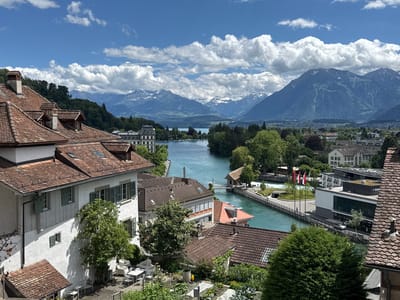 This high-angle, wide shot captures a scenic view of Thun, Switzerland, with the Aare River flowing through the city towards a distant lake, framed by snow-capped mountains under a bright, cloudy sky. In the foreground, the terracotta-tiled roofs and white stucco walls of traditional Swiss architecture dominate the left side of the frame, with several trees and outdoor seating arrangements visible. As the view extends toward the river, a modern building with a green roof stands on the right bank, adorned with Swiss and possibly cantonal flags. The river itself is a vibrant turquoise, flanked by green banks and further structures, including a picturesque building with a red roof and a small waterfall or rapid area. The background is a panorama of lush green hills transitioning into the majestic, snow-dusted peaks of the Alps. The scene is bathed in bright sunlight, casting distinct shadows and highlighting the contrast between the urban elements and the natural landscape.