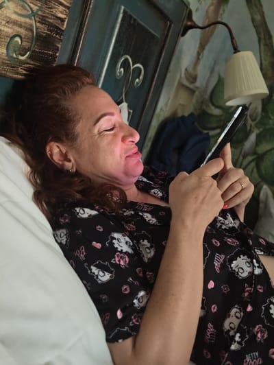 A woman with red hair and earrings is lying in bed, looking at her phone. She is wearing a black pajama top with a Betty Boop pattern and pink roses. Her left arm is resting on a white pillow, and her right hand is holding the phone. The background shows a wall with a mural and a lamp. The image is taken from a high angle. The location is the city of Pozzuoli, Italy.