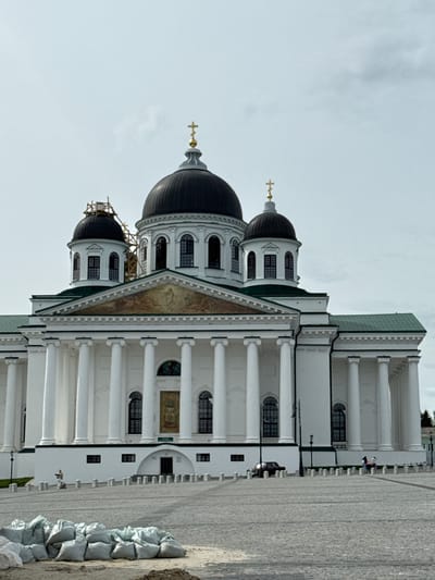 The image shows the Sobornaya Square in Arzamas, Russia, featuring the Resurrection Cathedral. The cathedral, a white building with black domes and golden crosses, dominates the scene. Its architecture is characterized by columns and arched windows. Some scaffolding is present on one of the domes, indicating ongoing maintenance. A section of the paved square is visible, as are sandbags along the edge. A few people are seen in the background. The sky is overcast, suggesting a cloudy day.