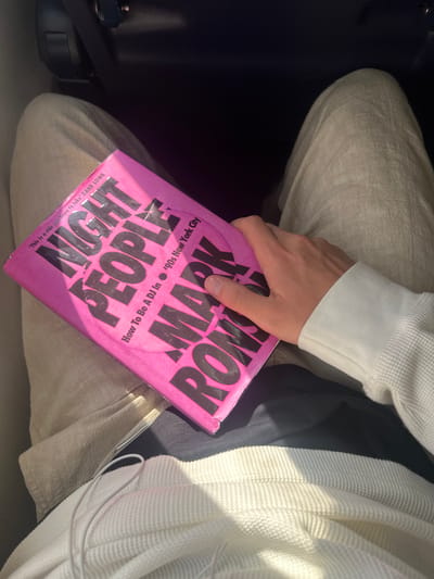 A person is holding a pink book in their lap. The book is titled "NIGHT PEOPLE: How To Be A DJ In '90s New York City" by MARK ROMV. The person is wearing light-colored pants and a white long-sleeved shirt. The book is held in the person's left hand. The setting appears to be indoors. The location is not within Braintree, United Kingdom, as the book title refers to New York City.