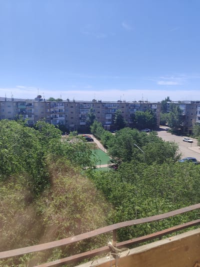 This image depicts a view of a residential area in the city of Abay, Kazakhstan, on a clear, sunny day. In the foreground, a rusty metal railing and a wooden surface suggest a balcony or elevated viewpoint. Beyond this, dense green trees dominate the middle ground, obscuring much of the lower portion of the buildings.
A multi-story apartment building, characteristic of Soviet-era architecture, is visible behind the trees. The building is made of concrete and grey material, with numerous windows and balconies. Several satellite dishes are mounted on the roof. Below the apartment building, a paved courtyard area is visible, with cars parked and a green sports court or playground area with some play equipment. The sky is a bright, cloudless blue, indicating fair weather. The overall impression is a typical urban residential landscape in Kazakhstan during summertime.