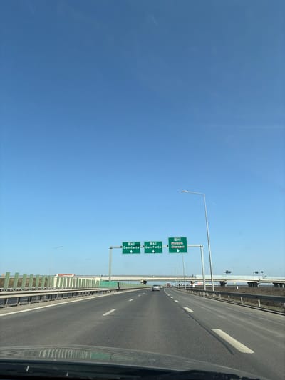 The image depicts a multi-lane highway under a clear, bright blue sky. The viewpoint is from inside a vehicle, with the top of a dashboard visible at the bottom of the frame. The highway features three lanes traveling forward, marked with white dashed and solid lines, and a paved shoulder on the right.
Overhead, a green gantry supports three road signs. The two signs on the left indicate "A2 Constanta" with a downward arrow, designating two lanes for that destination. The sign on the far right indicates "A0 Ploiești Urziceni" with a downward arrow, for the adjacent lane.
Along both sides of the highway, numerous silver lampposts are visible. To the left, a green sound barrier or fence runs parallel to the road. In the mid-distance, a concrete overpass or bridge spans across the highway. Several vehicles, primarily passenger cars, are visible in the distance traveling in the same direction. Another bridge or overpass structure is visible further in the background to the right. The surrounding environment appears to be relatively flat with sparse, dry vegetation on the verges.
Based on the road signs indicating "A2 Constanta" and "A0 Ploiești Urziceni," the scene is located on the A2 motorway in Romania, specifically at or approaching the interchange with the Bucharest Ring Road (A0) on the eastern side of Bucharest, in the vicinity of Glina, Ilfov County, Romania.