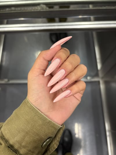 The image shows a person's hand with long, light pink, pointed nails. The nails are manicured and polished, with a subtle shimmer. The hand is wearing a green jacket, and the person's other hand is visible in the background. The scene is likely captured in a private setting, possibly a home or a beauty salon, based on the appearance of the environment. The coordinates provided correspond to a location in Ramla, Israel.