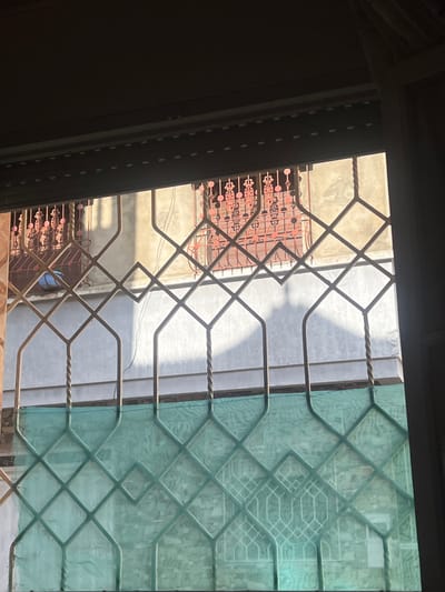 The image depicts a view from inside a structure in Ksar-el-Kebir, Morocco, looking outwards through a window onto an opposing building. The foreground shows a partially raised dark roller shutter at the top, beneath which is a window secured by light-colored, geometric metal grilles with twisted bar sections. The lower half of this window is covered by a translucent, light green mesh. Across from this viewpoint, the midground features the facade of another building, constructed of light-colored, possibly stuccoed, material showing weathering. This building has at least two visible windows, each equipped with ornate reddish-brown metal grilles displaying intricate scroll or floral patterns. Strong, directional sunlight illuminates sections of this opposing building, casting prominent shadows, including a large, irregular shadow on its central wall. A blue bucket or container is visible near the lower-left window of the opposing building. No people are present, and no actions or interactions are depicted. The environment suggests a dense urban or residential area.