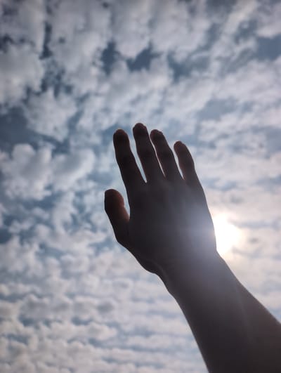 A hand is raised towards a cloudy sky, with the sun peeking through the clouds in the upper right corner of the image. The image is taken from a low angle, looking up. The clouds are white and fluffy, and the sky is a light blue. The hand is the only subject in the image. The image is of good quality with no visible artifacts. It is difficult to determine the exact location in Varengeville-sur-Mer, France from the image alone, but it is likely an outdoor location.