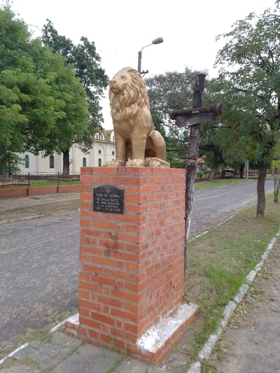 The image shows a golden lion statue atop a red brick pillar in Villa Hayes, Paraguay. The pillar bears a plaque commemorating the "Club de Leones de Villa Hayes 25 años sirviendo a la comunidad 1983-29.09.2008."  A weathered wooden cross stands adjacent to the pillar. A paved road runs alongside the statue, with trees lining either side. In the background, a white building with arched windows can be seen.  A street light is visible behind the statue.