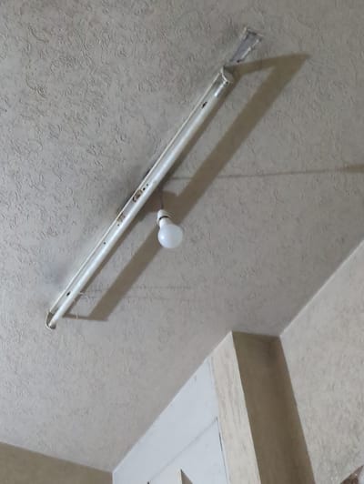 The image shows a close-up, angled view of a textured ceiling in what appears to be an indoor setting. The main object in focus is a long, white fluorescent light fixture, partially detached from the ceiling at one end, causing it to hang down. The fixture casts a distinct shadow on the ceiling. Above and slightly to the right of the fixture, a single, round, white light bulb is visible. This bulb is also casting a shadow. Part of a ceiling cornice or architectural feature is visible in the upper right quadrant of the image. The textured surface of the ceiling is a neutral, light color, possibly beige or off-white, with a raised, swirled pattern. The lighting suggests it is daytime, with artificial light from the bulb creating sharp shadows. There is no text visible in the image. The scene appears to be in a residential or commercial building.