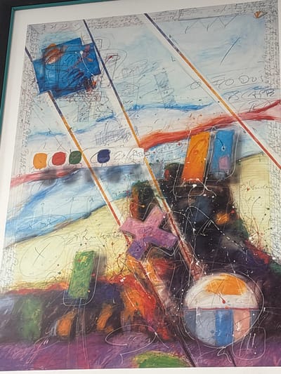 This is a framed abstract painting with a white mat and frame. The artwork features bold, energetic lines and splatters in a variety of colors, including blues, oranges, reds, purples, and greens. Geometric shapes and abstract forms are interwoven throughout the composition, with some appearing layered or transparent. There are also calligraphic scribbles and what appear to be notes or text scattered across the surface.

Notable details include a large circular motif divided into orange and blue sections, a purple cross-like shape, and several rectangular blocks of color. The background includes areas of light blue and yellow, overlaid with darker, more intense colors. The overall impression is one of dynamism and complexity.

Visible text at the bottom of the painting appears to be a signature and date. Partially legible words include "Mülheim a.d. Ruhr" and "1983". It is possible the text refers to the artist and location of creation.