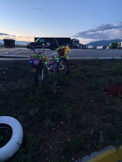 The image depicts an outdoor scene at what appears to be a roadside or truck stop in Barakovo, Bulgaria, during dusk or dawn, given the soft, diffused light and the colors in the sky.
In the foreground and midground, on a grassy and dirt verge adjacent to a paved area, two children's-sized bicycles are prominently displayed. Both bikes are painted in shades of purple and pink and are extensively decorated. They feature string lights, some emitting blue light and others white or yellow, wrapped around their frames and handlebars. Artificial yellow and white flowers are also intertwined with the bikes, with one bike having a small potted plant on its handlebars. A large, green, succulent-like plant grows in the grass nearby. To the lower right, a white-painted tire lies on the ground. A red wagon or wheelbarrow is partially visible in the upper right corner of the grassy area.
On the paved road or parking lot to the left, several semi-trucks are visible, some parked and others potentially in motion in the distance. These include green, white, and a dark-colored truck. Closer to the foreground on the left, a dark SUV or car is parked, and a black rectangular container or bin sits on the ground next to it. White text "KHM..." or "KHMU..." is partially visible on the side of this dark vehicle. The road features a clear yellow demarcation line.
The background shows distant mountains under a light blue sky with a prominent, elongated cloud formation stretching across. No specific activity is occurring; the scene is static, with the decorated bicycles suggesting a memorial or a permanent decorative display. The overall impression is one of peacefulness at the close or start of a day.
