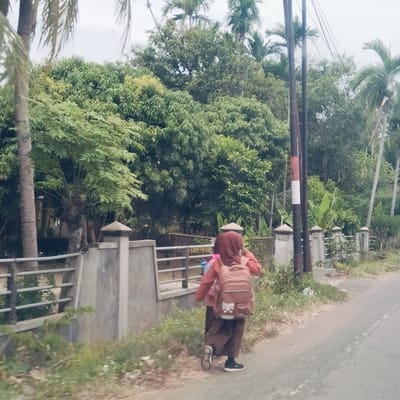 The image depicts two individuals walking along the side of a paved road in a verdant, rural or semi-rural area, consistent with Padangkasab, Indonesia. The scene is illuminated by diffused daylight under an overcast sky, casting no harsh shadows.
The prominent subject, viewed from behind, is wearing a reddish-brown hijab and a matching long-sleeved top over dark, loose-fitting trousers or a long skirt, paired with black sneakers. They are carrying a sizable light brown backpack. Another person, partially visible next to them, also appears to be carrying a backpack and walking in the same direction.
To the left of the road, there is a low concrete fence topped with decorative pillars and metal railings. Beyond the fence, the landscape is dense with lush green foliage, featuring numerous tropical trees including tall palm trees and other leafy trees with what appear to be developing fruits.
On the right side of the road, there is a narrow dirt shoulder with grass and weeds. A dark utility pole with a distinctive red and white striped section stands further down the road, with power lines stretching across the upper frame. The road surface is grey asphalt with faint white markings along its edge. The overall impression is a tranquil, everyday scene in a tropical environment. No decipherable text is clearly visible.
