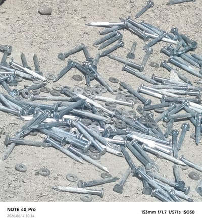 A pile of metallic nails and washers is scattered across a gravel surface. The nails are of varying lengths and are oriented in different directions, with some appearing to be partially embedded in the ground. Several circular washers are also visible amongst the nails. The scene is illuminated by natural light, casting subtle shadows on the ground. The overall impression is one of construction materials left in an outdoor setting. The image was taken in Kyzyl-Yulduz, Uzbekistan.