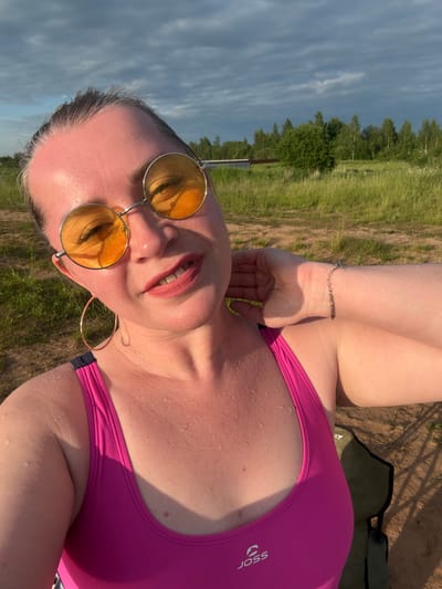 A woman with grey-streaked brown hair pulled back is taking a selfie outdoors in Sapunov Bor, Russia. She is wearing large, round, yellow-tinted sunglasses and a bright pink one-piece swimsuit. Her head is tilted to her left, and her right hand is touching the back of her neck. She has large hoop earrings on her left ear. Small droplets of water are visible on her skin and swimsuit. A thin chain bracelet with small beads and a charm is on her right wrist. In the background, there is a grassy field, trees, and a cloudy sky. A green folding chair is partially visible to the right of the frame.
