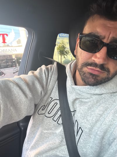 A man wearing sunglasses and a grey hoodie with black lettering is seated in a car, fastened with a seatbelt. The view through the car window shows a street scene in Mesa Geitonia, Cyprus, with visible signs and storefronts. A building with the name "COBIANA ELECTRONIC CIGARETTES" is visible with a logo with red "T" and white wings.