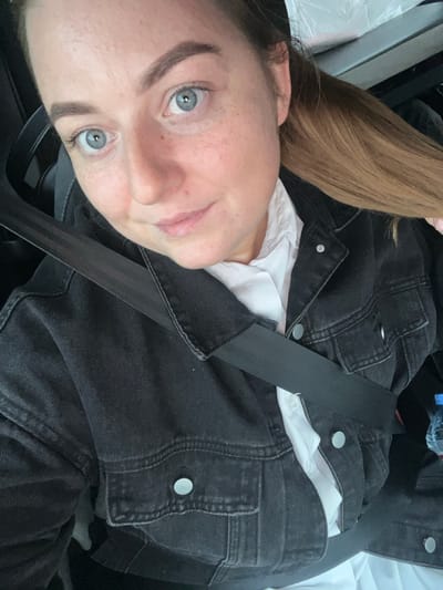 A young woman with fair skin, light blue eyes, and light brown hair is shown in a close-up selfie. She is wearing a white top underneath a black denim jacket. A black seatbelt is fastened across her chest. The background is indistinct, but appears to be inside a vehicle. The location cannot be determined from the image.