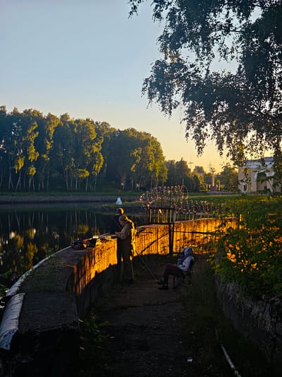 The image depicts an outdoor scene at sunset, possibly in a rural or park-like setting. Two people are present, both engaged in fishing. One person, wearing a light-colored jacket and pants, stands near a concrete embankment, holding a fishing rod. Another person is seated on a folding chair, also holding a fishing rod.
The setting features a calm body of water on the left, reflecting the surrounding trees and the sky. A dense line of trees with lush green foliage borders the water. On the right side, a concrete wall runs along the embankment, illuminated by the warm glow of the setting sun. The ground leading to the embankment is covered in dirt and grass. In the background, there are hints of industrial structures, possibly power lines or a small building, with a bright yellow-orange sky suggesting the end of the day.
The time of day is clearly indicated by the low angle of the sun and the warm, golden light bathing the scene. The weather appears clear and calm, conducive to fishing. The overall mood is peaceful and serene, with the individuals focused on their activity. No text is visible in the image. The location context provided is "Водник Снт, Russia."