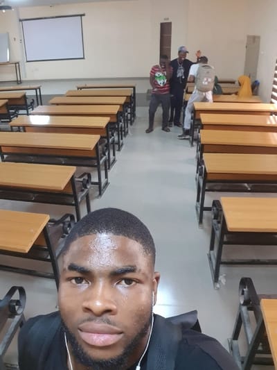 This image depicts the interior of a classroom or lecture hall in Panhauya, Nigeria. The room features light-colored walls and a light-colored floor. On the front wall, a large white projection screen is mounted, with a portion of a whiteboard visible to its left. Numerous two-person student desks, constructed with light-colored wooden tops and black metal frames, are arranged in parallel rows, extending from the foreground into the background.
In the immediate foreground, a young adult male with a dark complexion, short dark hair, and a short beard is positioned, facing the camera directly. He is wearing a dark-colored shirt and white in-ear headphones. His forehead appears slightly shiny. He is taking a selfie.
In the midground, three young adult males are standing between rows of desks. From left to right: The first male, dark-skinned, wears a red, black, and white striped shirt and dark pants; he is laughing with his hands near his face. The second male, dark-skinned, wears a dark long-sleeved top and a light-colored baseball cap worn backward; his right arm is raised. The third male wears a light-colored shirt, light-colored pants, and a khaki backpack; his back is mostly turned, but his head is slightly turned towards the others, and his right arm is also raised. This group appears to be engaged in a social interaction.
In the far right background, a person is seated at a desk, partially obscured. This individual wears a yellow head covering and light-colored clothing. A window with horizontal blinds is visible on the right wall.