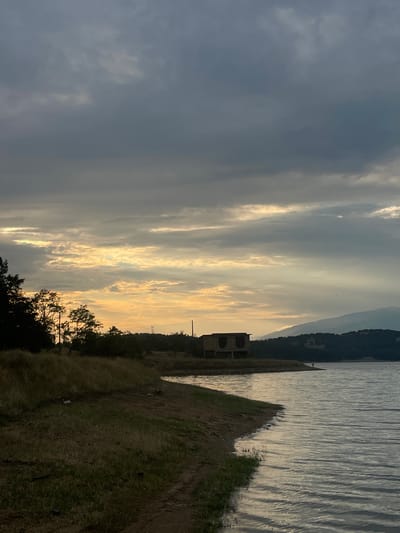 This is a vertical, eye-level, full shot of a serene lakeside landscape at dusk. The sky is dramatic, with dark, heavy clouds dominating the left side, interspersed with bright, golden streaks of sunlight breaking through on the right, creating a stark contrast. In the distance, the silhouette of a forested mountain range forms the horizon.
The foreground features a grassy embankment sloping down towards a calm body of water. The water reflects the muted colors of the sky, with subtle ripples suggesting a gentle breeze. On the embankment, several trees with dark foliage are visible, some appearing as silhouettes against the lighter sky. A portion of an old, stone building with arched openings stands on the right side of the frame, adding a touch of historical intrigue to the scene. The ground is covered in dry, yellowed grass, hinting at the end of summer or beginning of autumn.
The overall mood is peaceful and contemplative, amplified by the soft, fading light of sunset. The presence of the building suggests a past human presence, now reclaimed by nature. The lack of any visible people or explicit activity points towards a quiet, natural moment. The time of day is clearly evening, with the dramatic lighting and cloud formations characteristic of sunset. No text is visible in the image. The location is identified as Domlyan, Bulgaria, which aligns with the natural landscape depicted.