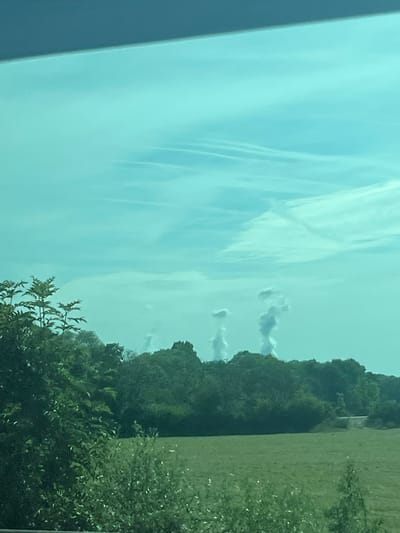 A landscape scene shows vibrant green trees and foliage on the right side of the frame, contrasted against a sky filled with wispy clouds and contrails on the left. The sky transitions from a darker blue to a lighter hue closer to the horizon. Overall, the image captures a sense of natural beauty and expansive space.