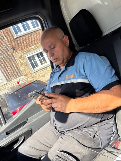 The image shows a man lying down inside a vehicle, with his eyes closed, appearing to be resting. He is wearing a blue shirt with orange lettering that reads "DYNA" and dark-colored pants. His arm is resting on his chest, and he is holding a smartphone in his hand, with the screen facing upwards. The interior of the vehicle is visible, including a black seat and a black door panel. Outside the vehicle, through the window, a brick building with white-framed windows can be seen, suggesting an outdoor setting. The lighting suggests it is daytime, with sunlight casting shadows inside the vehicle. The overall impression is one of a brief moment of rest or relaxation during a journey or work break. The location context provided is Henri-Chapelle, Belgium.