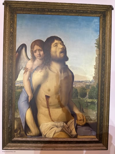 The painting depicts a scene with two central figures. The primary figure is a man, likely representing Christ, with his eyes closed and mouth open, exhibiting signs of suffering. He is depicted shirtless, revealing wounds, notably a prominent wound on his torso with dripping blood. A shroud is draped around his lower body. To his left, a winged figure, an angel, is positioned behind him, partially obscured by the man's body. The angel has a sorrowful expression and is holding the man. The background shows a landscape with trees, rocky formations, and a distant cityscape with architectural structures. The painting is housed in a decorative frame. The lower left corner of the image contains text that reads "© Museo Nacional del Prado - 2019". There is no indication of the city of Mazarrón, Spain, in the image or its associated text.