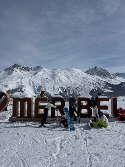 The image displays six individuals on a snow-covered slope at the Méribel ski resort, located within the Three Valleys ski area in France, accessible from the commune of Saint-Martin-de-Belleville. A large, three-dimensional "I ❤️ MÉRIBEL!" sign, constructed from dark-colored material, stands prominently in the midground.
Five individuals are positioned around the sign. From left to right:
1. A person standing on skis, wearing a white jacket and dark pants, leaning against the "M" of "MÉRIBEL" while holding ski poles.
2. A person kneeling in the snow in front of the "É", dressed in dark attire, with a blue "Burton" brand snowboard propped upright beside them.
3. A person standing on skis between the "É" and "R" letters, wearing a dark top and white pants, holding ski poles.
4. A person standing on skis between the "R" and "I" letters, wearing a black top and white pants, with their right arm raised in a gesture.
5. A person seated on the snow to the right of the "L", wearing a white jacket and bright neon yellow-green pants, with skis on their feet.
A sixth individual, dressed in red and black ski gear, is partially visible further to the right in the background. All individuals are wearing winter outerwear, including jackets, pants, and headwear or helmets with goggles. The background features extensive snow-covered alpine mountains under a sky with scattered light clouds. Ski tracks are visible on the foreground slope.