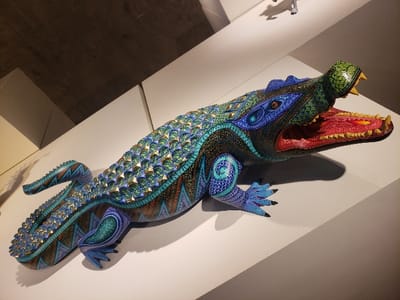 A brightly painted wooden alligator statue sits on a white platform. The alligator is decorated with patterns of teal, blue, green, purple, orange, gold, and brown. The statue's mouth is open, revealing a bright red interior and yellow teeth. The alligator has claws on its feet and its tail curls to the left. The background consists of a slanted white wall and a grey concrete ceiling.